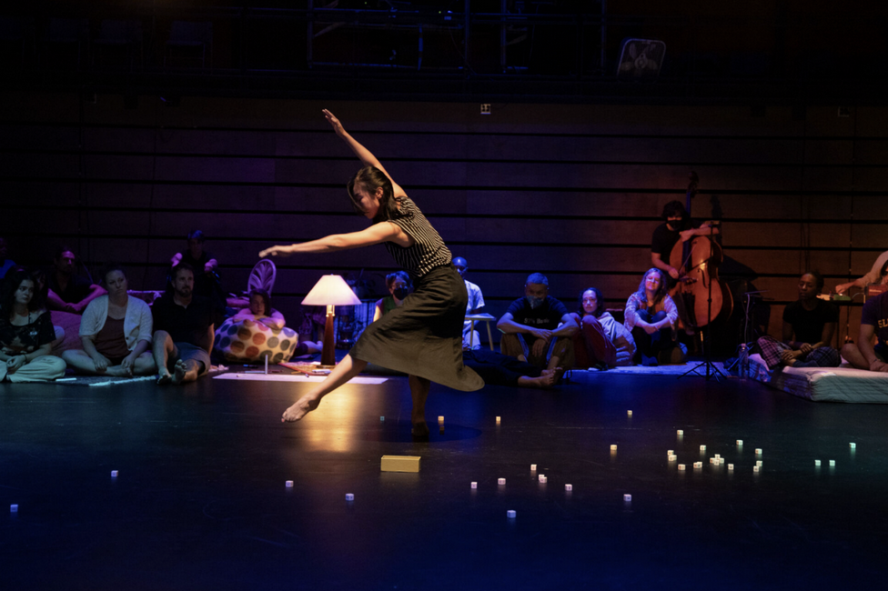 Dancer performing on darkened stage with performers observing in background